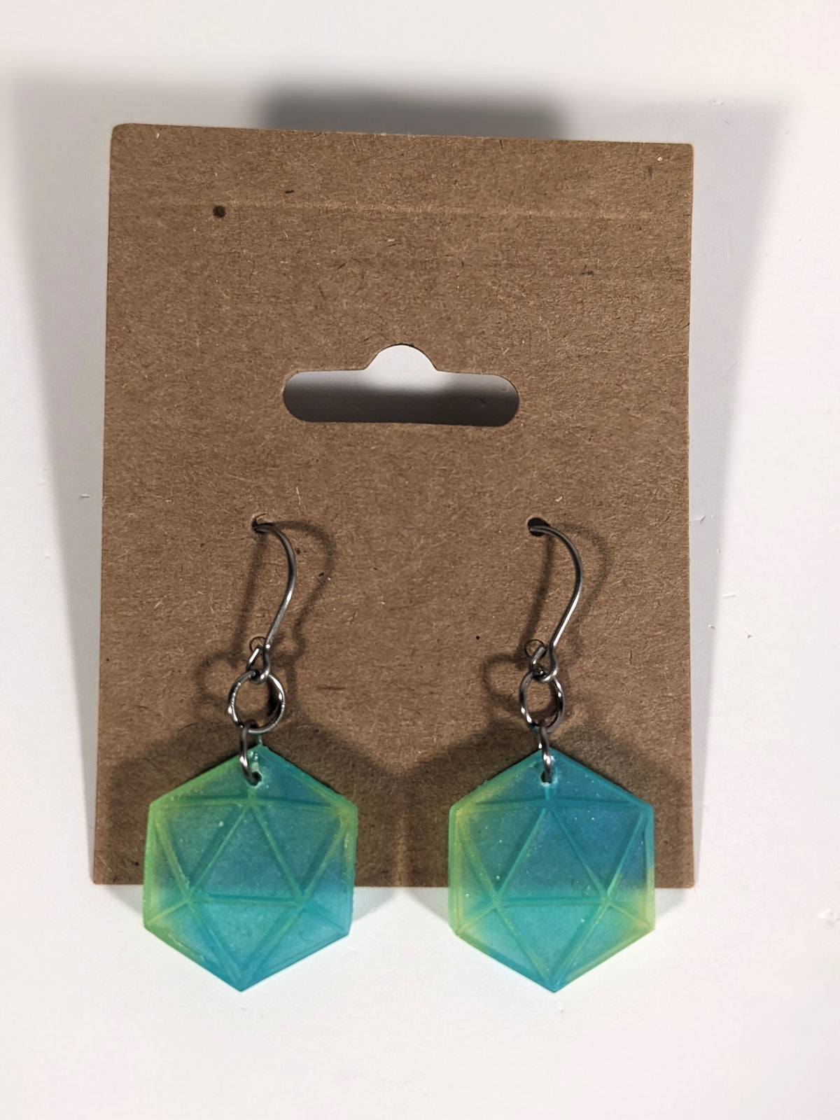 A pair of teal and yellow dangly, flat d20 earrings with silver color titanium and niobium hardware on a kraft earring card on a white background.