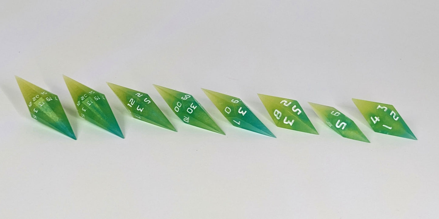A mid angle photo of a complete set of handmade diamond DnD dice that are yellow and teal swirls with white numbers arranged in a straight line with bottom points angled slightly forwards and towards the right. They are lined up with the 2 d20s on the left going down to the d4 on the right side. The highest value number of each die is facing forward.