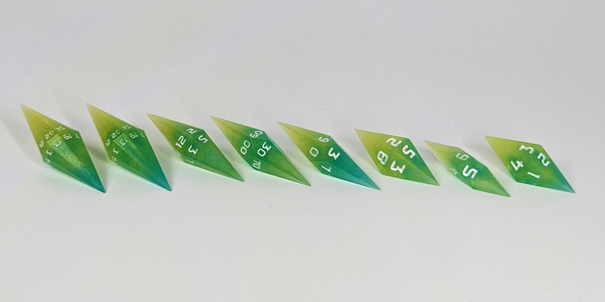 A mid angle photo of a complete set of handmade diamond DnD dice that are yellow and teal swirls with white numbers arranged in a straight line with bottom points angled slightly forwards and towards the right. They are lined up with the 2 d20s on the left going down to the d4 on the right side. The highest value number of each die is facing forward.