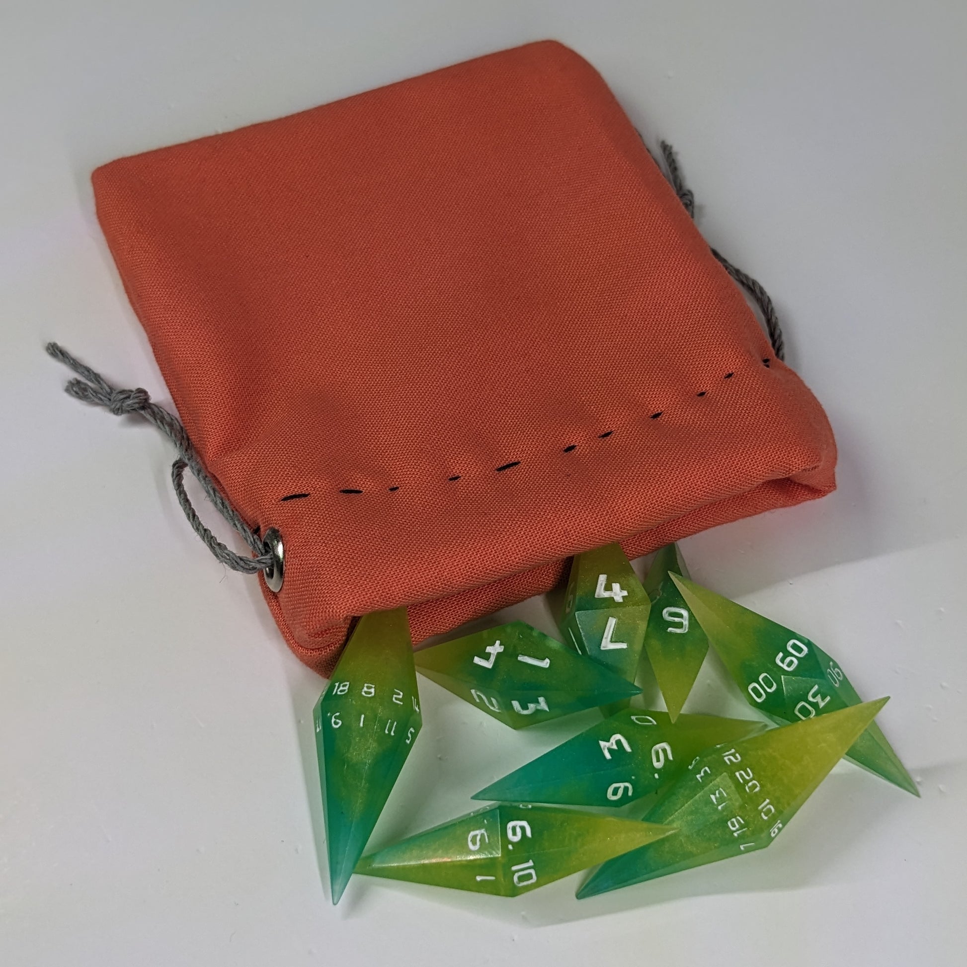 Yellow and teal swirls with white numbers diamond handmade dice spilling out of an orange drawstring bag on a white background. The set features 2 d20s and matching resin dice, showcasing the unique shape of these artisan dice.