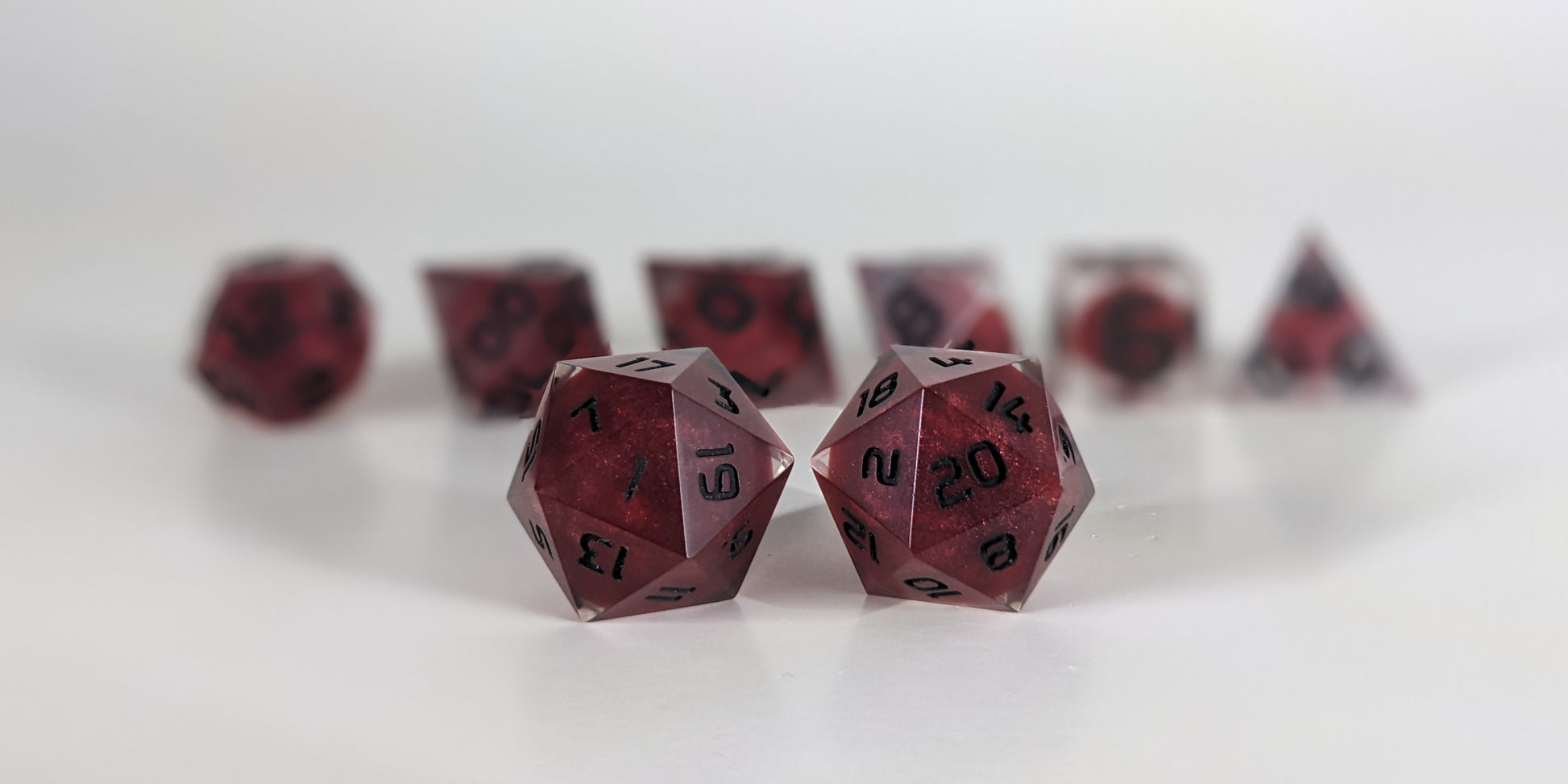 A straight on photo of a standard set of DnD dice that are clear with red, spherical inserts and black numbers focusing on 2 d20s in the center. The d20 on the left has the 1 face centered and the d20 on the right has the 20 face centered. The other 6 dice are lined up in the background but are out of focus, while the d20s are in sharp focus.