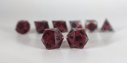 A straight on photo of a standard set of DnD dice that are clear with red, spherical inserts and black numbers focusing on 2 d20s in the center. The d20 on the left has the 1 face centered and the d20 on the right has the 20 face centered. The other 6 dice are lined up in the background but are out of focus, while the d20s are in sharp focus.