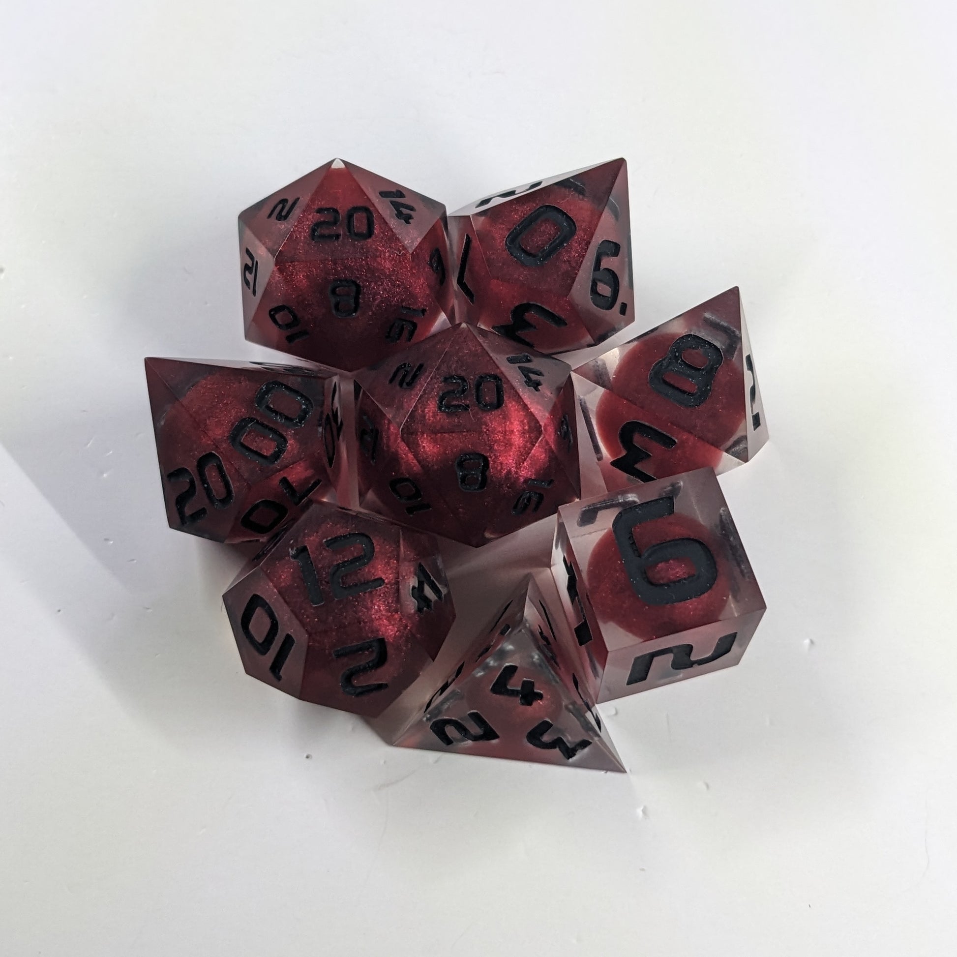 A complete set of 8 handmade artisan dice that are clear with red, spherical inserts and black numbers. The DnD dice set is arranged in a circle and features 2 D20s, one of which is in the center surrounded by the other 7 dice.