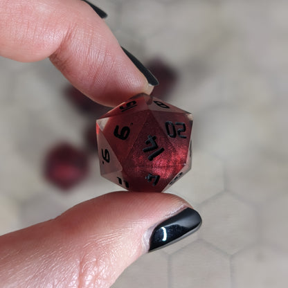 The d20 die is being held up to the camera to show off the red, spherical insert and the dark glow the black numbers give in the clear shell. The 14 face is most angled toward the camera.