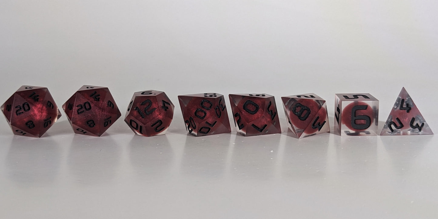 A straight on photo of a complete set of polyhedral dice that are clear with red, spherical inserts and black numbers arranged in a straight line. They are lined up with the 2 d20s on the left going down to the d4 on the right side. The highest value number of each die is facing forward.