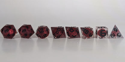 A straight on photo of a complete set of polyhedral dice that are clear with red, spherical inserts and black numbers arranged in a straight line. They are lined up with the 2 d20s on the left going down to the d4 on the right side. The highest value number of each die is facing forward.
