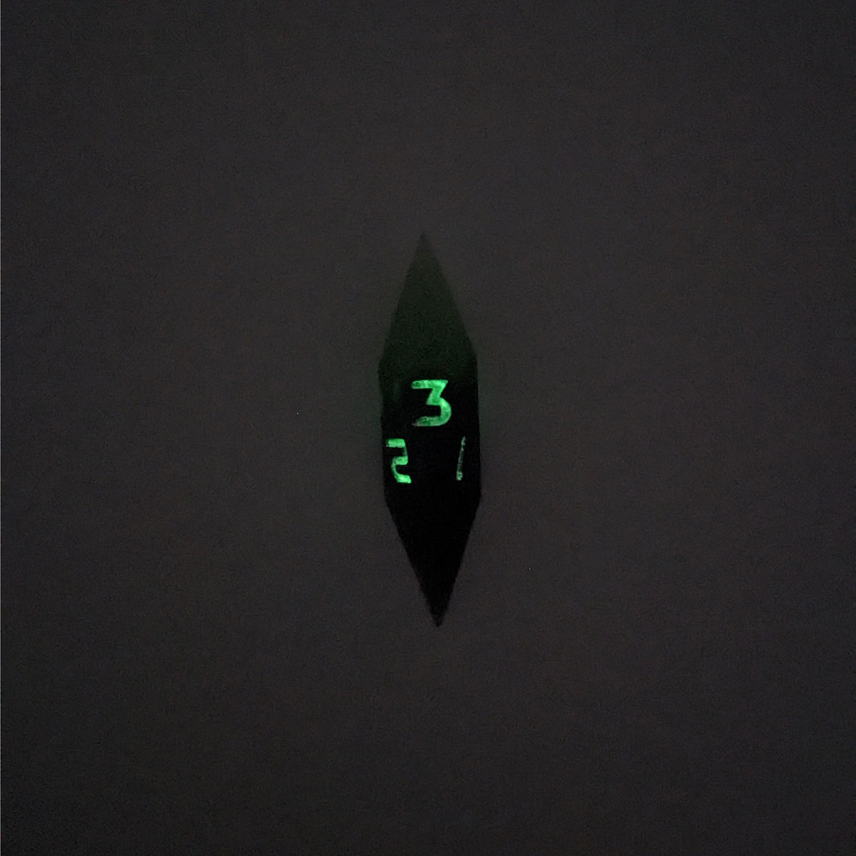 A high angle view of a diamond d3 with swirls of black and green with purple numbers that glow in the dark on a white background. This photo is taken in the dark to show the purple numbers glowing green in the dark.