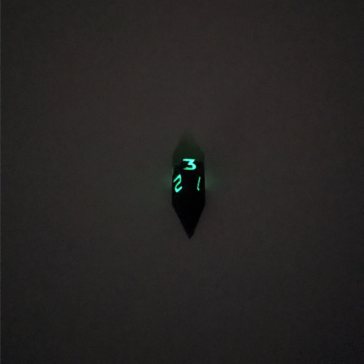 A mid angle view of a diamond d3 with swirls of black and green with purple numbers that glow in the dark on a white background. This photo was taken in the dark to show the purple numbers glowing green in the dark.