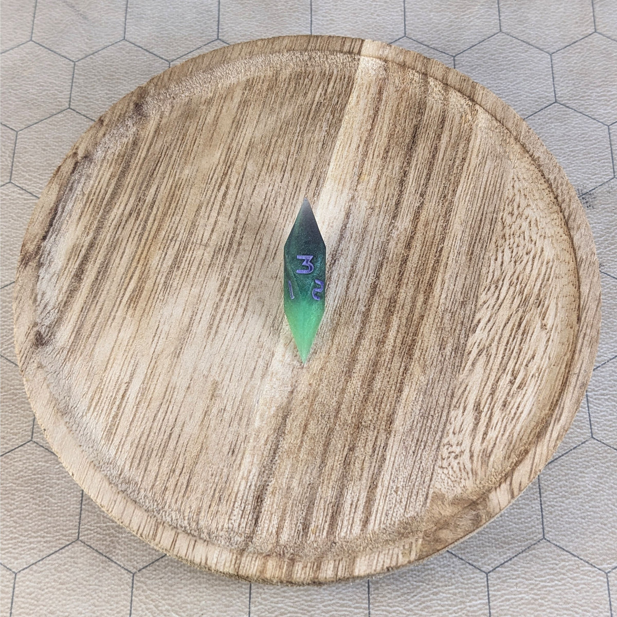 A high angle view of a diamond d3 with swirls of black and green with purple numbers that glow in the dark. It is sitting in the center of a wooden circle.