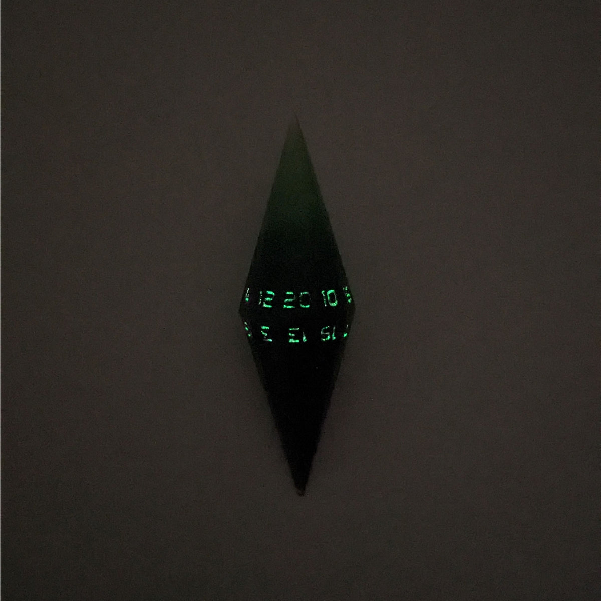 A high angle view of a diamond d20 with swirls of black and green with purple numbers that glow in the dark on a white background. This photo is taken in the dark to show the purple numbers glowing green in the dark.