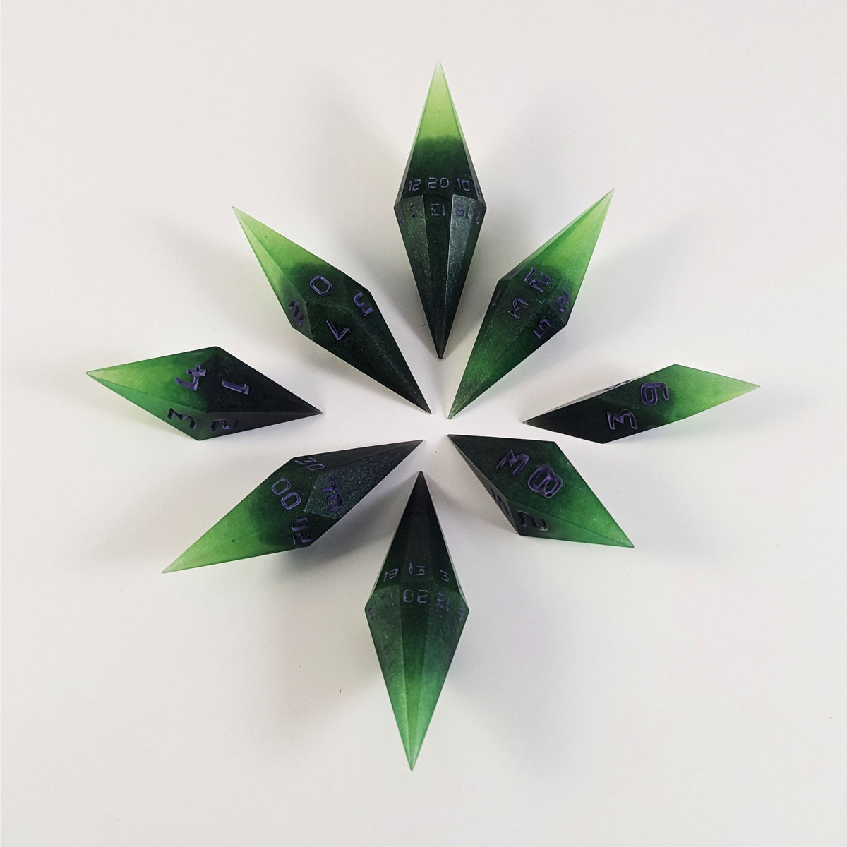A complete set of diamond shaped resin dice featuring 2 d20s. These handmade dice are black and green with purple numbers that glow in the dark and are clustered in a circle with the bottom points angled toward a central point on a white background.
