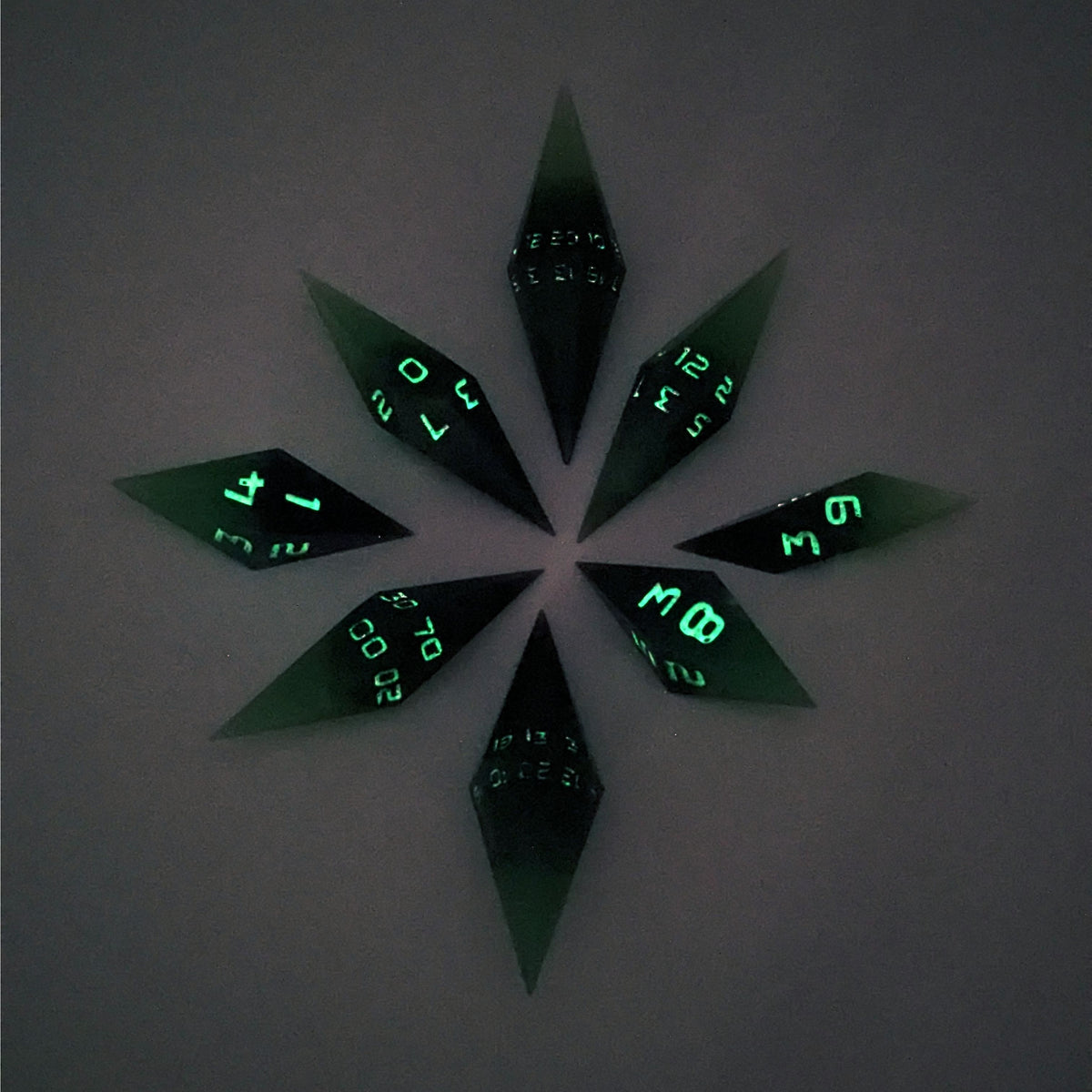 A complete set of diamond shaped resin dice featuring 2 d20s. These handmade dice are black and green with purple numbers that glow in the dark and are clustered in a circle with the bottom points angled toward a central point on a white background. This photo was taken in the dark to show the purple numbers glowing green.