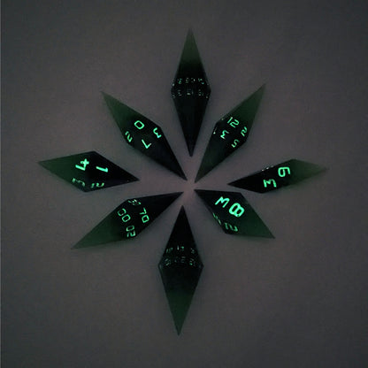 A complete set of diamond shaped resin dice featuring 2 d20s. These handmade dice are black and green with purple numbers that glow in the dark and are clustered in a circle with the bottom points angled toward a central point on a white background. This photo was taken in the dark to show the purple numbers glowing green.