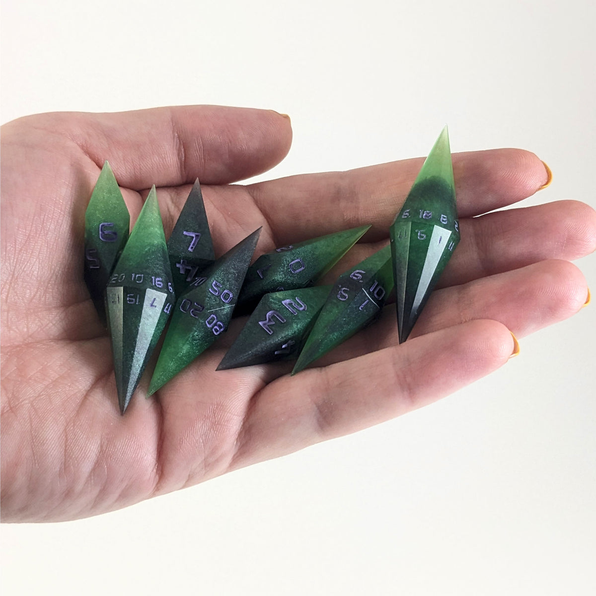 An 8 dice set of diamond style dice that are green and black with purple numbers that glow in the dark are held in a hand. The image shows that the set fits easily in a hand, making them overall smaller, though sharper, than a standard set of dnd dice.