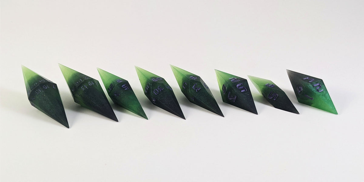 A mid angle photo of a complete set of handmade diamond DnD dice that are black and green with purple numbers that glow in the dark arranged in a straight line with bottom points angled slightly forwards and towards the right. They are lined up with the 2 d20s on the left going down to the d4 on the right side. The highest value number of each die is facing forward.