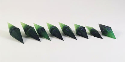 A mid angle photo of a complete set of handmade diamond DnD dice that are black and green with purple numbers that glow in the dark arranged in a straight line with bottom points angled slightly forwards and towards the right. They are lined up with the 2 d20s on the left going down to the d4 on the right side. The highest value number of each die is facing forward.