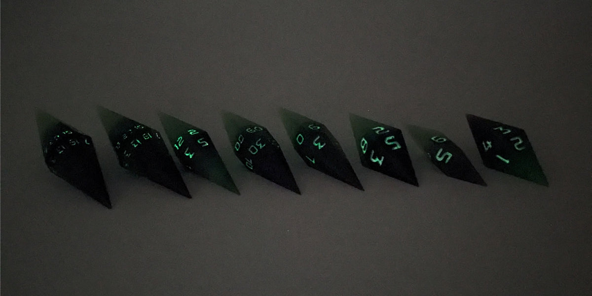 A mid angle photo of a complete set of handmade diamond DnD dice that are black and green with purple numbers that glow in the dark arranged in a straight line with bottom points angled slightly forwards and towards the right. They are lined up with the 2 d20s on the left going down to the d4 on the right side. The highest value number of each die is facing forward. The photo is taken in the dark to show the purple numbers glowing green.