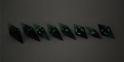 A mid angle photo of a complete set of handmade diamond DnD dice that are black and green with purple numbers that glow in the dark arranged in a straight line with bottom points angled slightly forwards and towards the right. They are lined up with the 2 d20s on the left going down to the d4 on the right side. The highest value number of each die is facing forward. The photo is taken in the dark to show the purple numbers glowing green.