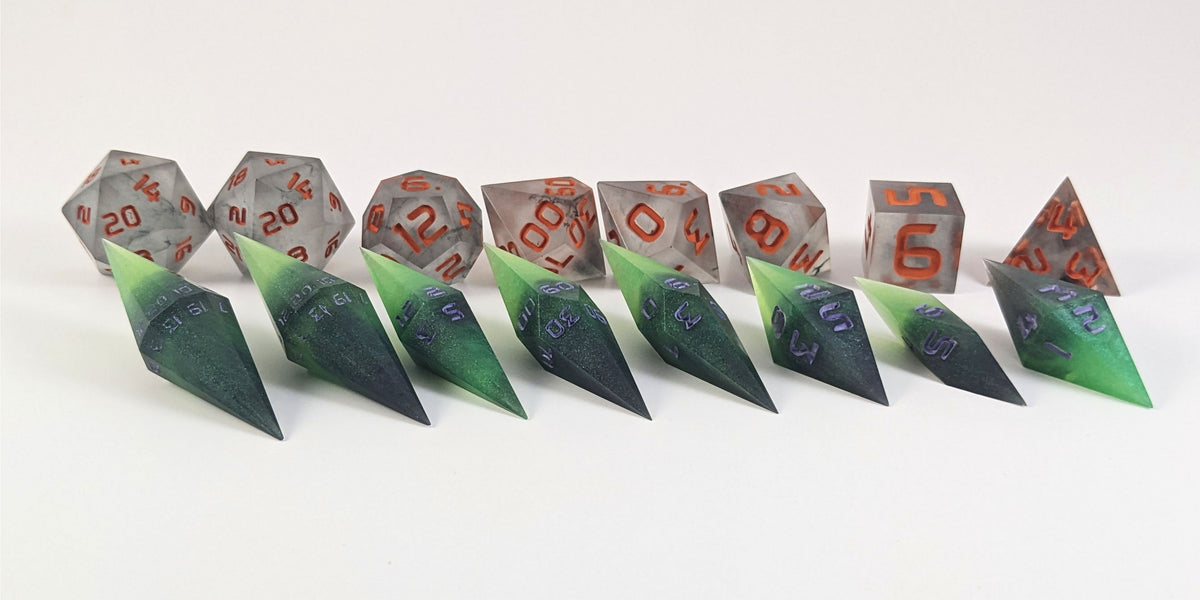A straight on photo of a complete set of dagger dice that are black and green with purple numbers that glow in the dark arranged in a straight line with points angled forward and to the right. They are lined up with the 2 d20s on the left going down to the d4 on the right side. Behind the dagger set is a standard set of polyhedral dice to allow for size comparison. The diamond dice are much smaller overall. The highest value number of each die is facing forward.