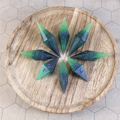 A set of handmade diamond dice that are black and green with purple numbers that glow in the dark arranged on a wooden circle. The resin dice have their tips facing outward, and the highest number of each die is facing up.