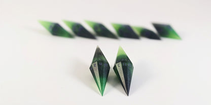 A straight on photo of a diamond set of DnD dice that are black and green with purple numbers that glow in the dark focusing on 2 d20s in the center. The d20 on the left has the 1 face up and the d20 on the right has the 20 face up. The other 6 dice are lined up in the background but are out of focus, while the d20s are in sharp focus.