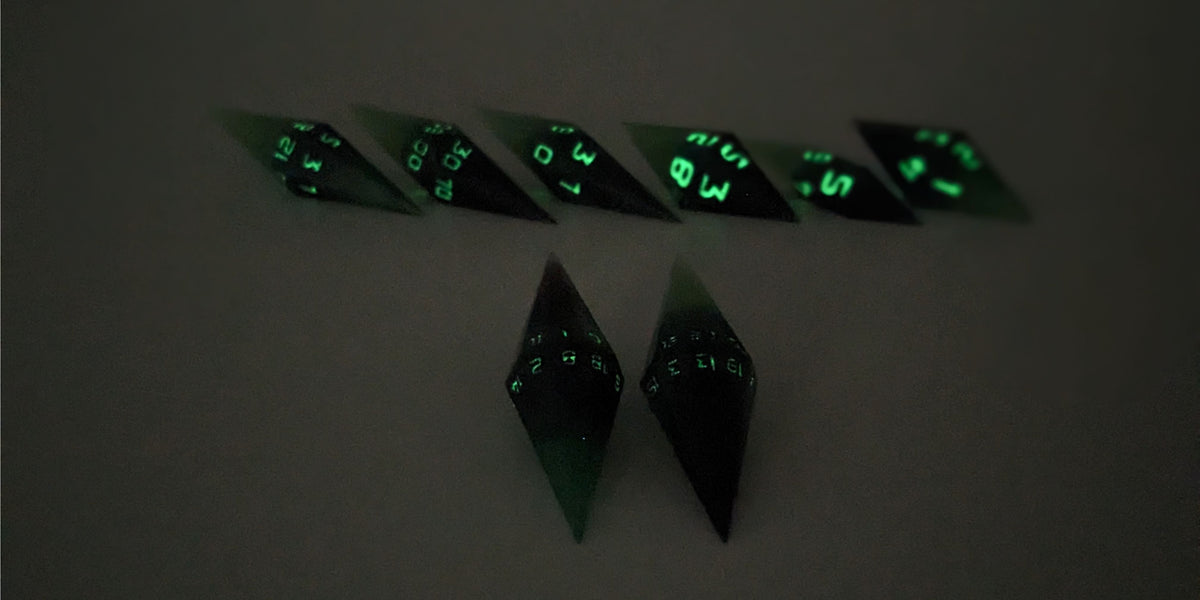 A straight on photo of a diamond set of DnD dice that are black and green with purple numbers that glow in the dark focusing on 2 d20s in the center. The d20 on the left has the 1 face up and the d20 on the right has the 20 face up. The other 6 dice are lined up in the background but are out of focus, while the d20s are in sharp focus. The photo is taken in the dark to show the purple numbers glowing green.