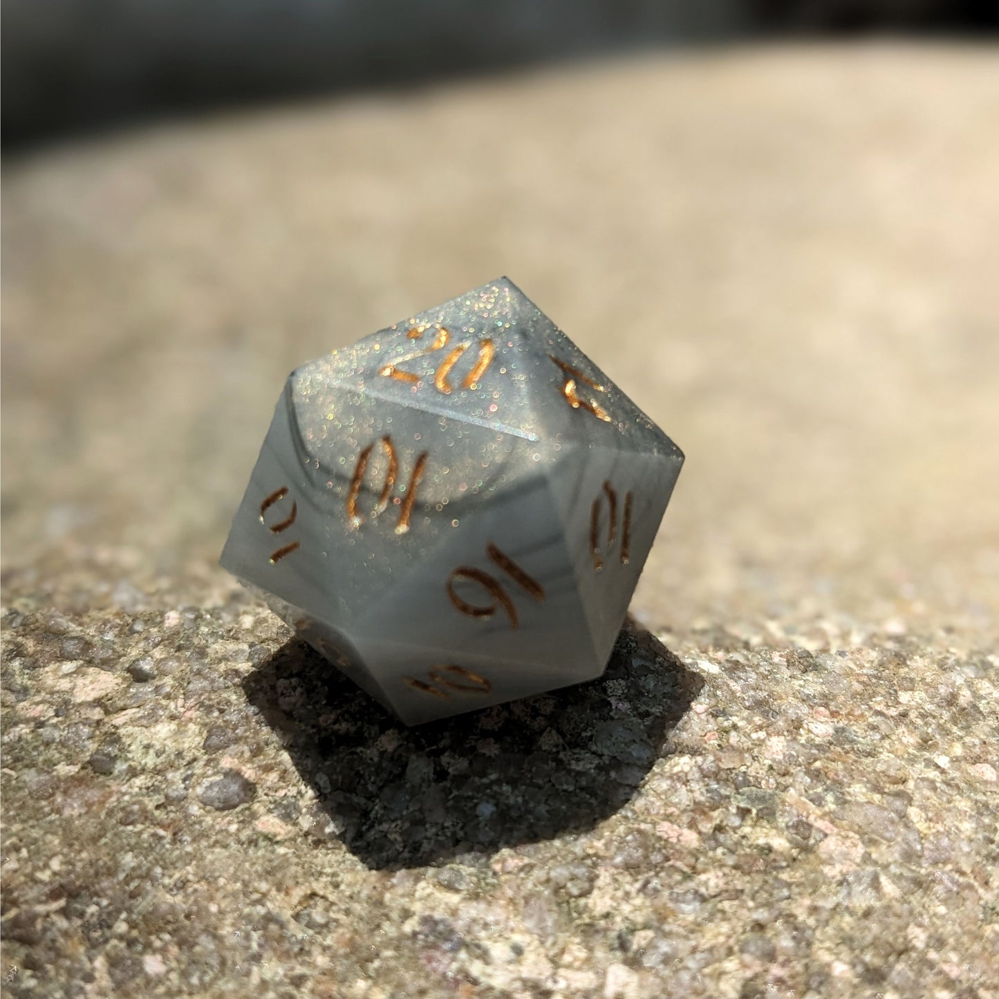 A close up photo of a black and white marbled with a slight gold shimmer reliable talent d20 with gold numbers sitting on a large rock. Faces that would be 9 or below are replaced with a 10.