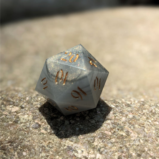 A close up photo of a black and white marbled with a slight gold shimmer reliable talent d20 with gold numbers sitting on a large rock. Faces that would be 9 or below are replaced with a 10.