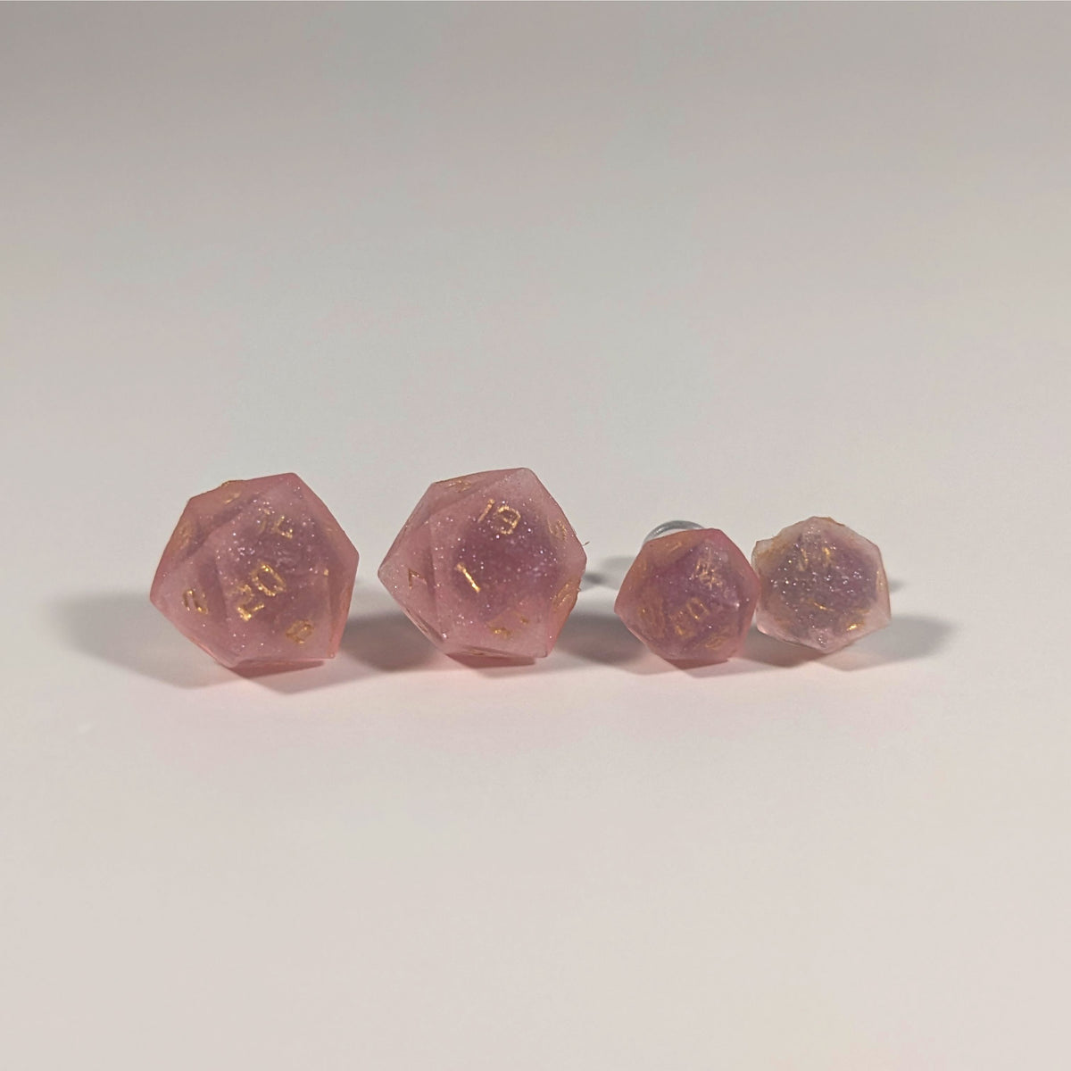 Two pairs of handmade d20 earrings with titanium hardware that are medium pink with gold numbers on a white background sit in a line with the larger pair on the left and the smaller pair on the right. The earrings on the left of each pair centers the 20 face and the earrings on the right of each pair centers the 1 face.