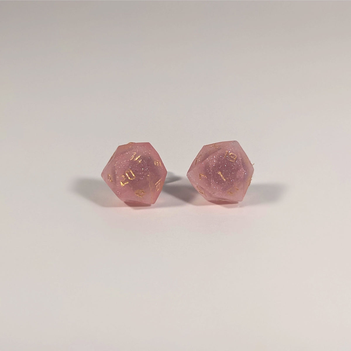 A single pair of the larger size handmade d20 earrings with titanium hardware that are medium pink with gold numbers on a white background. The earring on the left centers the 20 face and the earring on the right centers the 1 face.