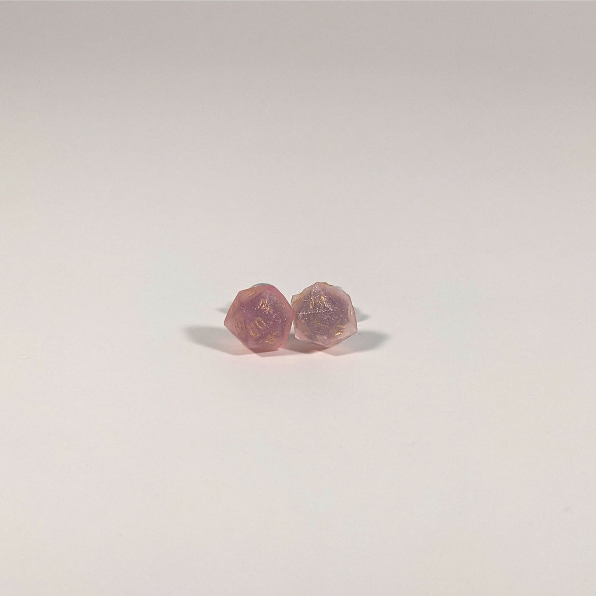A single pair of the smaller size handmade d20 earrings with titanium hardware that are medium pink with gold numbers on a white background. The earring on the left centers the 20 face and the earring on the right centers the 1 face.