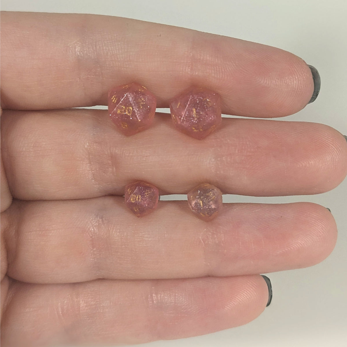 Two pairs of handmade d20 earrings with titanium hardware that are medium pink with gold numbers held between the fingers of a flat hand. The larger pair is between the pointer and middle finger, the smaller between the middle and ring finger. The earrings on the left of each pair centers the 20 face and the earrings on the right of each pair centers the 1 face.