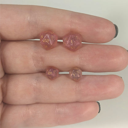 Two pairs of handmade d20 earrings with titanium hardware that are medium pink with gold numbers held between the fingers of a flat hand. The larger pair is between the pointer and middle finger, the smaller between the middle and ring finger. The earrings on the left of each pair centers the 20 face and the earrings on the right of each pair centers the 1 face.
