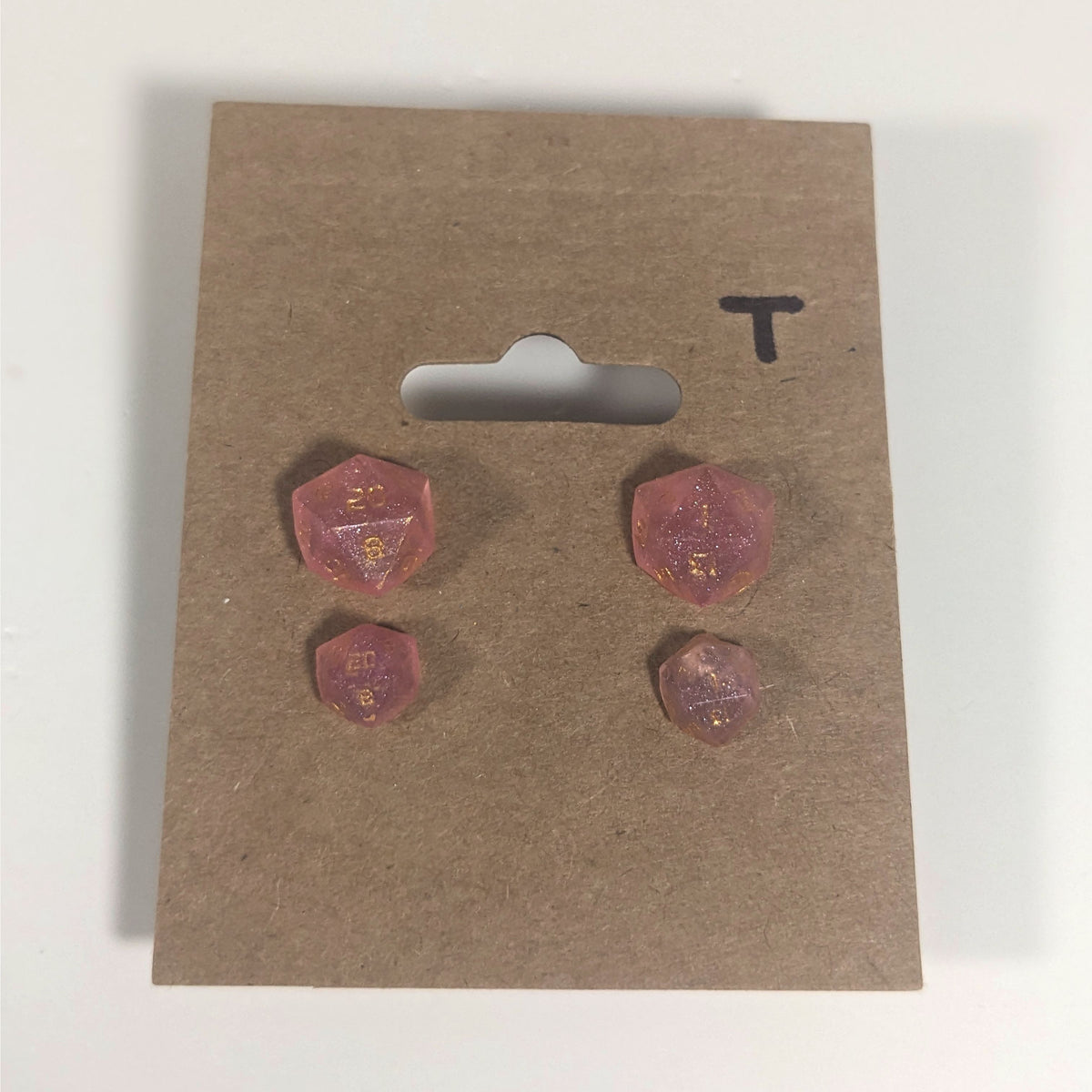 Two pairs of handmade d20 earrings with titanium hardware that are medium pink with gold numbers on a kraft brown earring card with a T in the top right corner. The earrings on the left of each pair centers the 20 face and the earrings on the right of each pair centers the 1 face.