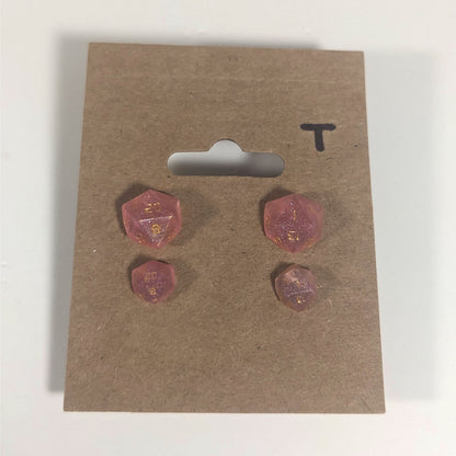 Two pairs of handmade d20 earrings with titanium hardware that are medium pink with gold numbers on a kraft brown earring card with a T in the top right corner. The earrings on the left of each pair centers the 20 face and the earrings on the right of each pair centers the 1 face.