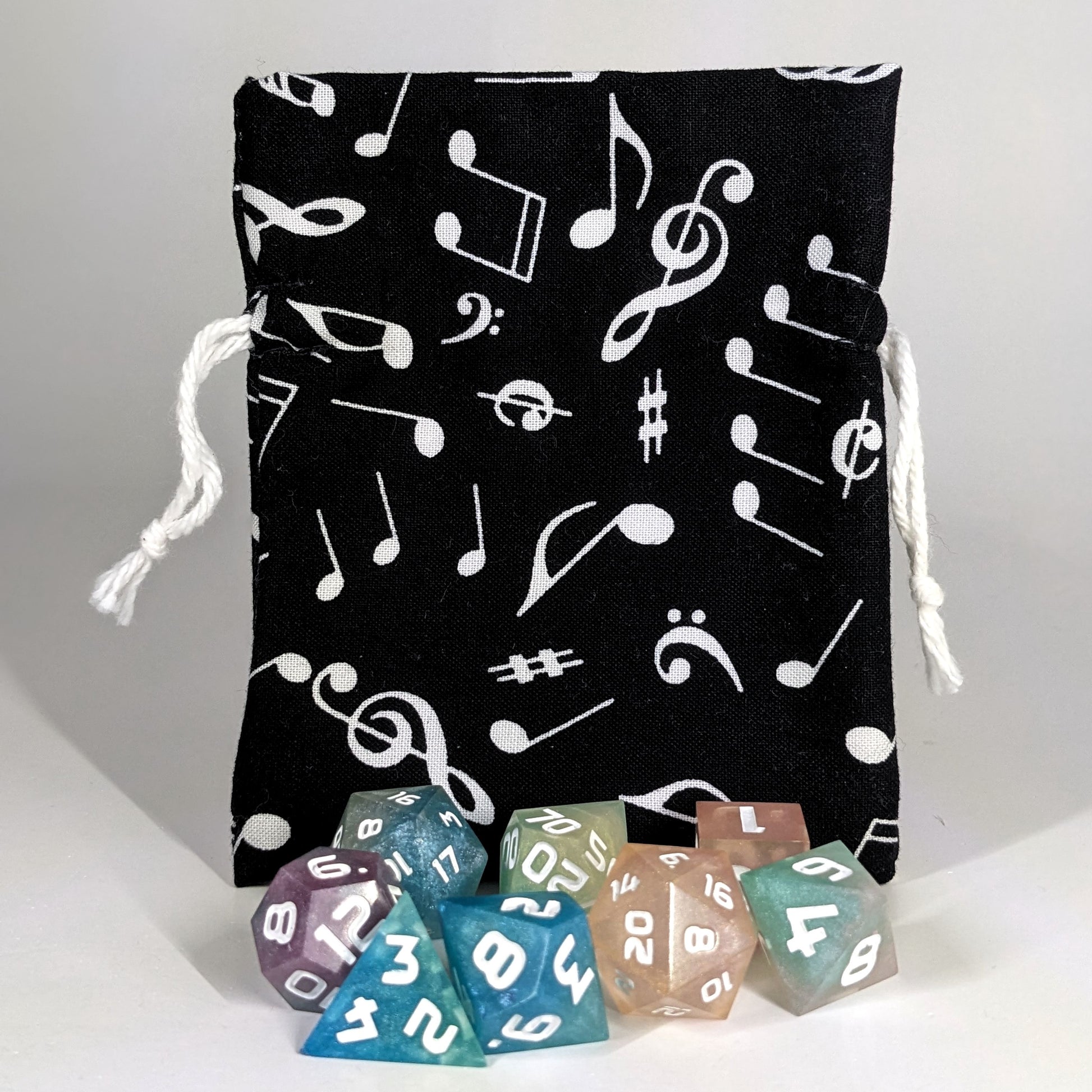 A small, handmade dice bag that is black with a white music notation pattern stands upright on a white background with a set of colorful dnd dice sitting in front of the dice.
