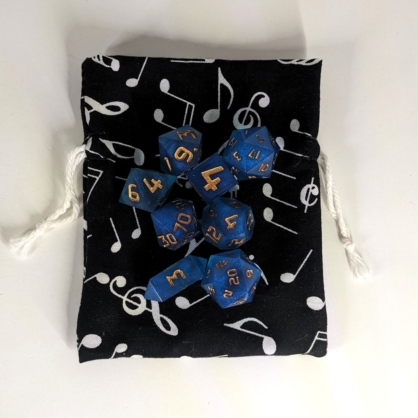 A small, handmade dice bag that is black with a white music notation pattern lays flat on a white background with a set of 8 blue, resin, polyhedral dice sitting on top.