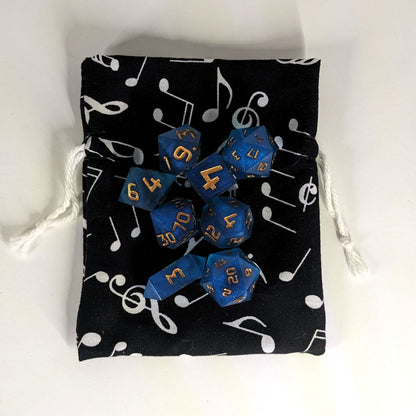 A small, handmade dice bag that is black with a white music notation pattern lays flat on a white background with a set of 8 blue, resin, polyhedral dice sitting on top.