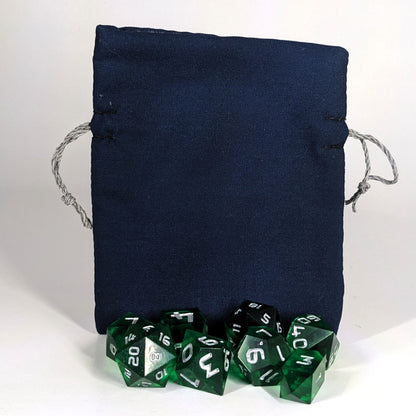 A small, handmade dice bag that is navy stands upright on a white background with a set of green dice with silver numbers sitting in front of it.
