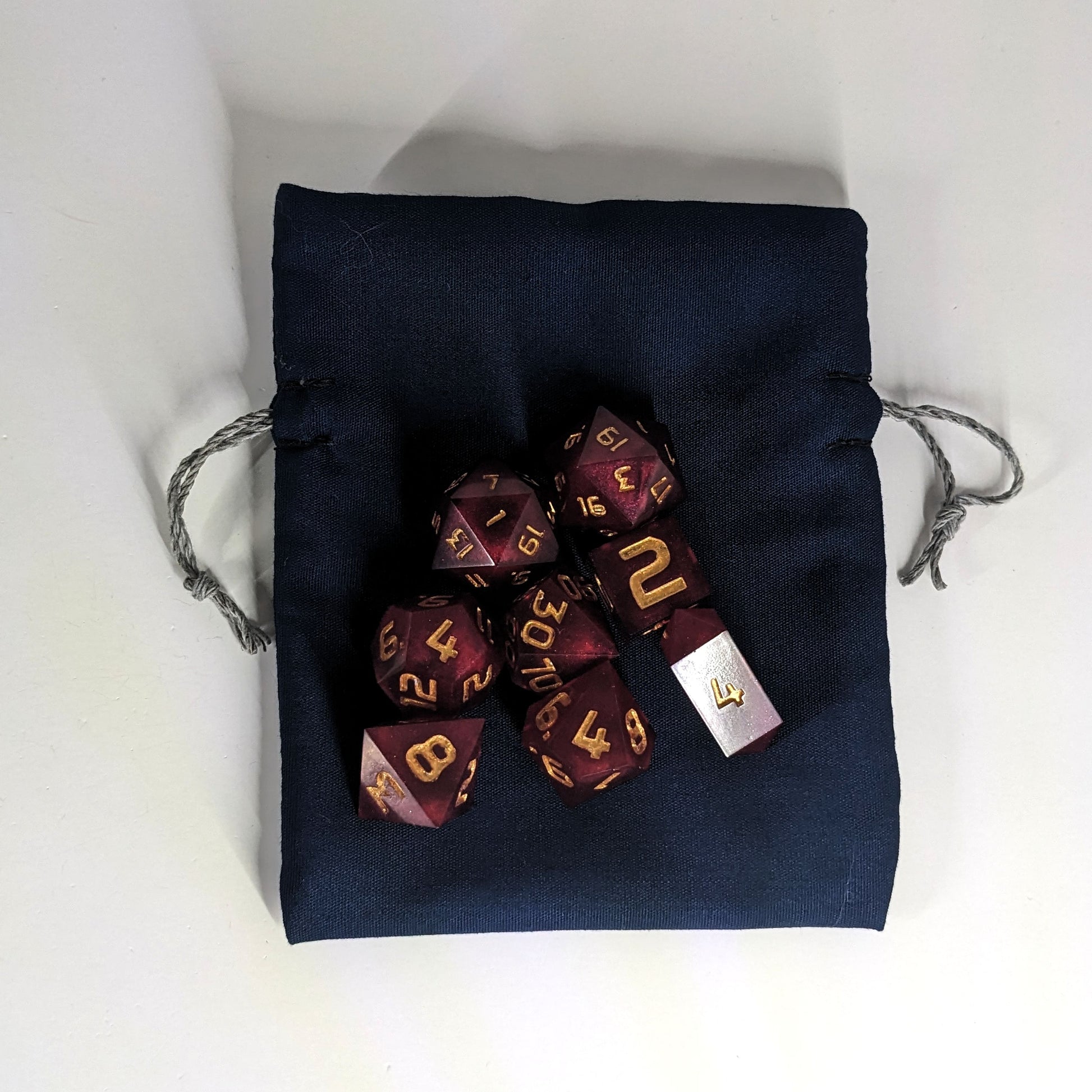 A small, handmade dice bag that is navy lays flat on a white background with a set of red dnd dice with gold numbers sitting on top.