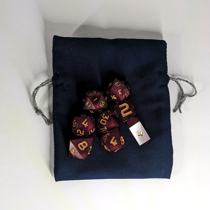 A small, handmade dice bag that is navy lays flat on a white background with a set of red dnd dice with gold numbers sitting on top.