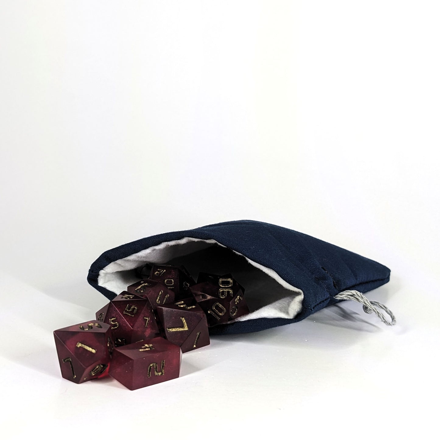 A small, handmade dice bag that is navy lays open with a set of red dnd dice with bronze numbers spilling out of it on a white background.