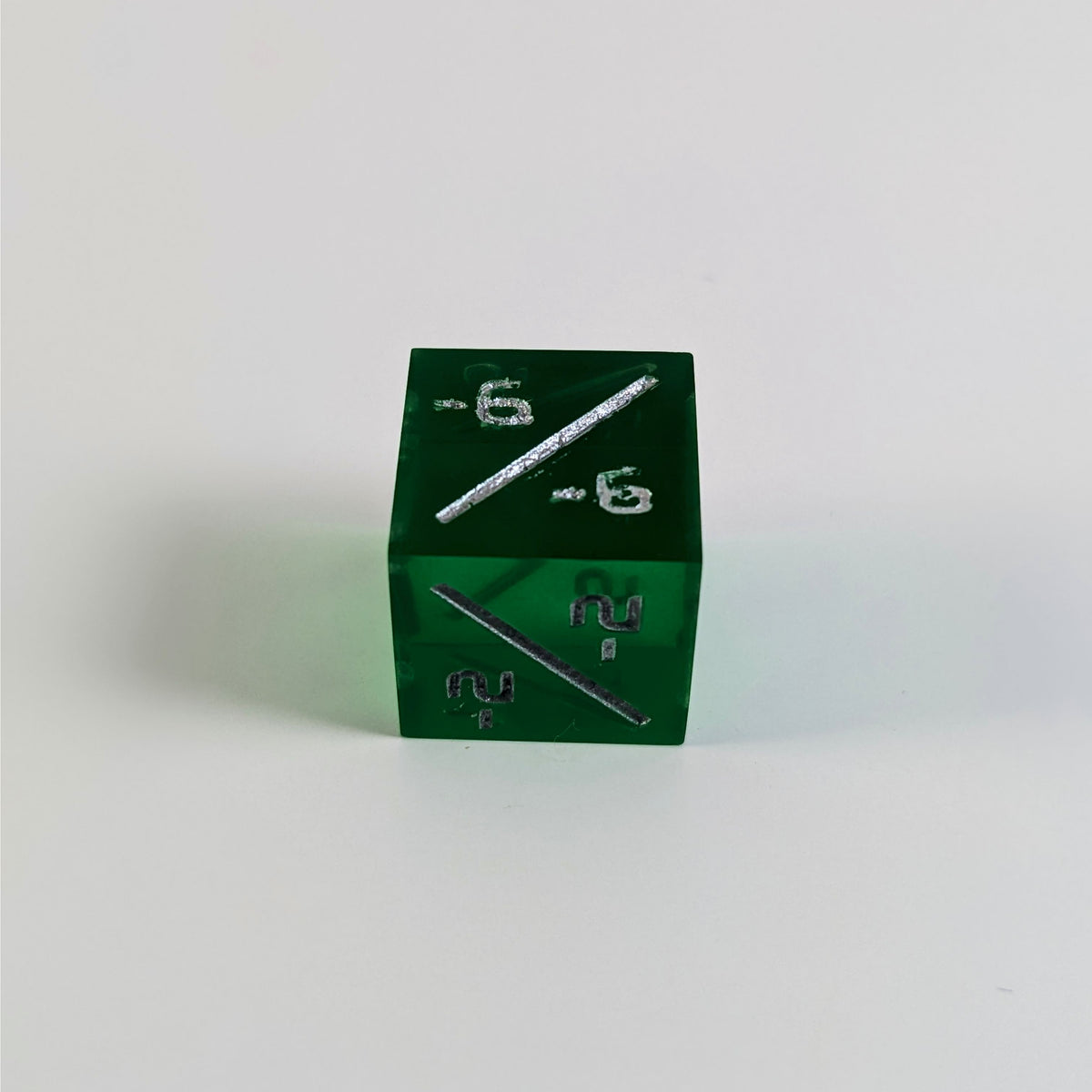 A low angle photo of a green negative counter d6 with silver numbers sits on a white background with the -6/-6 face up and the -2/-2 face pointed towards the camera.