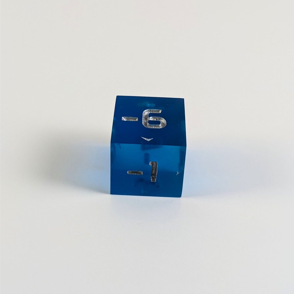 A low angle photo of a blue negative spindown d6 with silver numbers sits on a white background with the -6 face up and the -1 face pointed towards the camera.