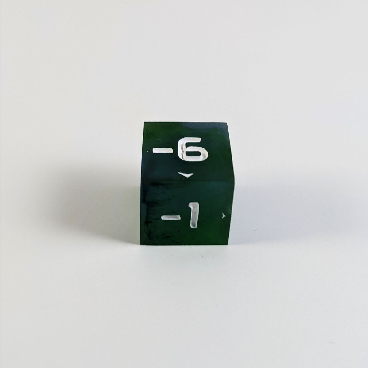 A low angle photo of a blue and green negative spindown d6 with white numbers sits on a white background with the -6 face up and the -1 face pointed towards the camera.