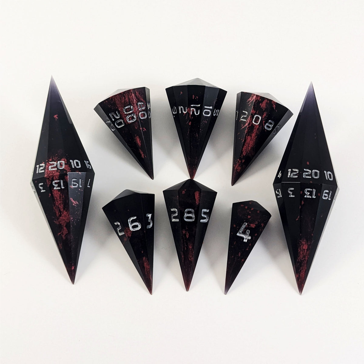 A dagger style DnD dice set that is black with crimson red splatter with silver numbers is arranged with a d20 each on the right and left, with the d%, d12, and d10 along the top and the d6, d8, and d4 along the bottom, all in line with the d20s.