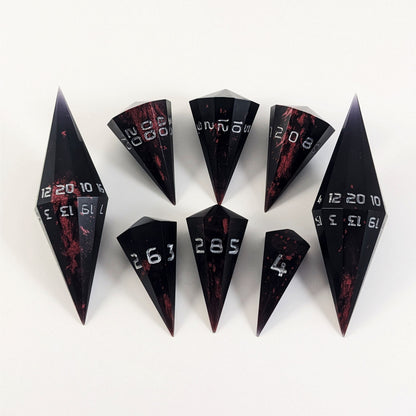 A dagger style DnD dice set that is black with crimson red splatter with silver numbers is arranged with a d20 each on the right and left, with the d%, d12, and d10 along the top and the d6, d8, and d4 along the bottom, all in line with the d20s.