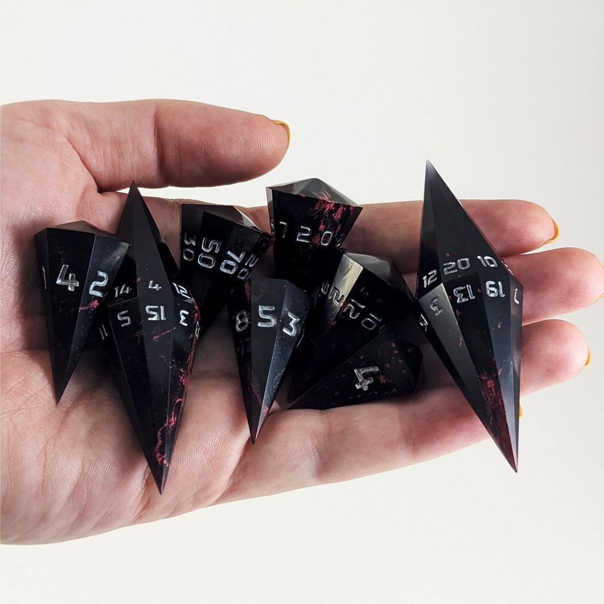 An 8 dice set of dagger style dice that are black with crimson red marbling with silver numbers are held in a hand. The image shows that the set just fits in a hand, making them overall larger than a standard set of dnd dice.