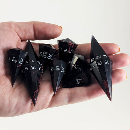 An 8 dice set of dagger style dice that are black with crimson red marbling with silver numbers are held in a hand. The image shows that the set just fits in a hand, making them overall larger than a standard set of dnd dice.