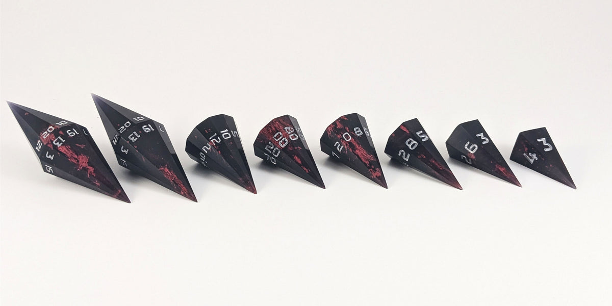 A straight on photo of a complete set of dagger DnD dice that are black with crimson red splatters with silver numbers arranged in a straight line with points angled toward the right. They are lined up with the 2 d20s on the left going down to the d4 on the right side. The highest value number of each die is facing forward.